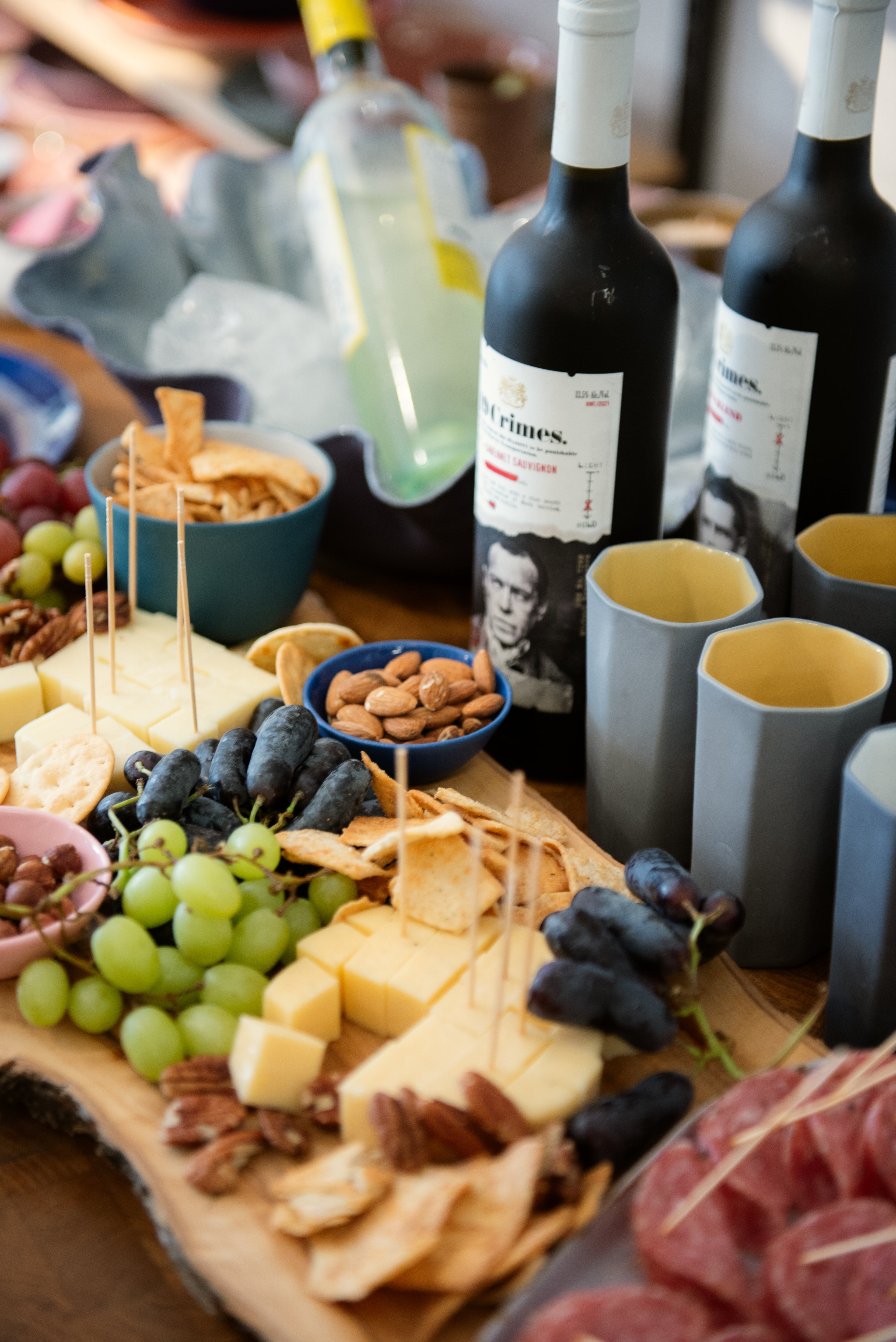 A charcuterie spread including various cheeses, meats, nuts, and grapes accompanied by ceramic mugs and plates on a table, indicative of a team-building pottery workshop event.